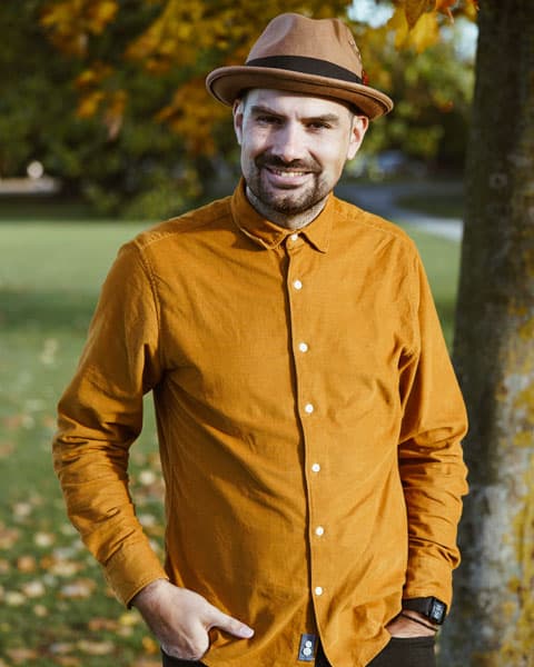 a white man in an orange shirt with a trilby hat standing next to a tree smiling 