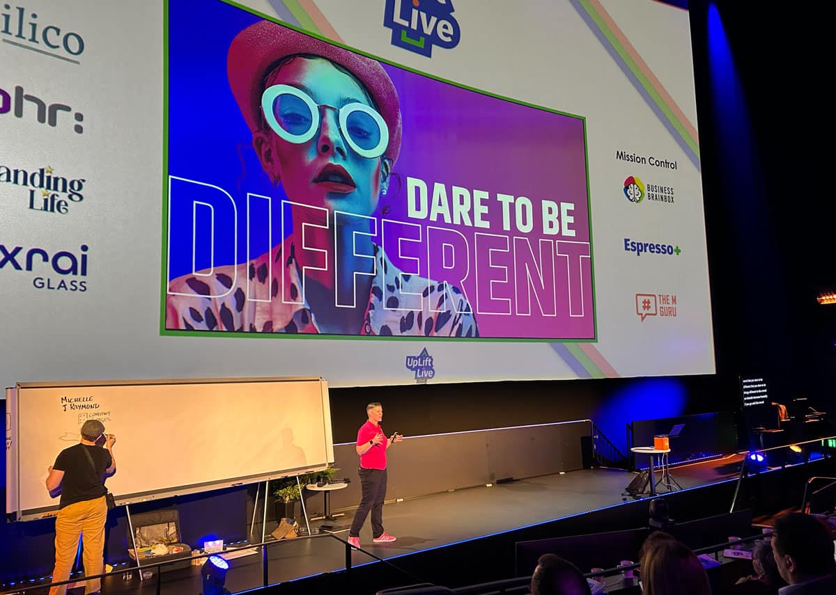 a white woman in a pink top speaking on stage with the slide behind her dare to be different.
